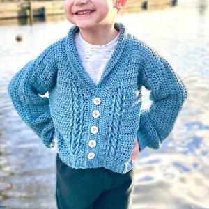 May include: A light blue crocheted cardigan with a cable knit pattern and white buttons. The cardigan has a v-neck and long sleeves.