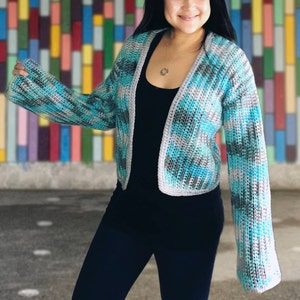 May include: A blue and gray crocheted cardigan with a long sleeve and open front. The cardigan is worn over a black top and black pants.