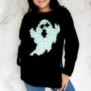May include: Black long-sleeve sweater featuring a crocheted ghost design in light green. The ghost has a black face and is centered on the front of the sweater. The sweater is paired with blue jeans.