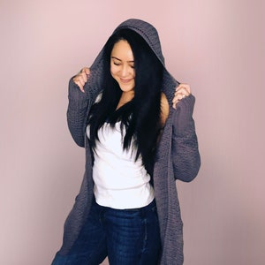 May include: A woman wearing a gray hooded cardigan over a white tank top and blue jeans.