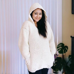 May include: A white crocheted hooded sweater with a loose fit and long sleeves. The sweater has a V-neck and pockets on the sides.
