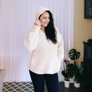 May include: A white crocheted hooded sweater with a loose fit and a textured pattern. The sweater is worn by a person with long dark hair.