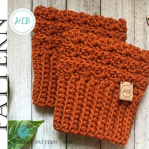 May include: Two crocheted orange cup cosies with a brown leather tag that says "HLB" on each. The cozies are on a white wooden surface.