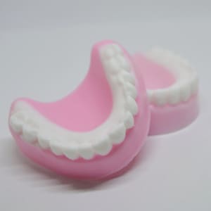 May include: Two pink plastic dentures with white teeth. The dentures are shaped like a mouth and are lying on a white surface.
