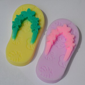 May include: Two pairs of flip-flop shaped soap bars. One is yellow with green floral accents and the other is purple with pink floral accents.