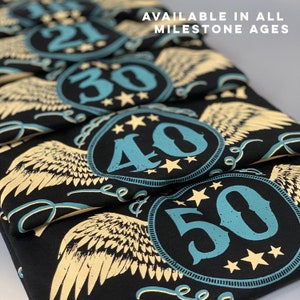 May include: A stack of black t-shirts with a teal number and gold wings design. The numbers on the shirts are 21, 30, 40, and 50. The text "AVAILABLE IN ALL MILESTONE AGES" is at the top of the image.