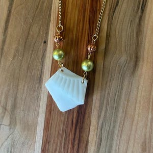 Puede incluir: Un collar de cadena dorada con un colgante blanco en forma de abanico. El collar presenta cuentas verde oliva y detalles cobrizos. El colgante es de color blanco roto claro y tiene una superficie texturizada.