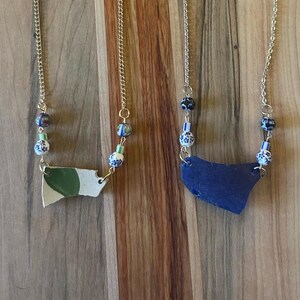 Può includere: Due collane con catene dorate e ciondoli unici. Un ciondolo è un frammento bianco con un disegno verde, l'altro è un frammento blu. Entrambe le collane hanno perline decorative blu, bianche e verdi.