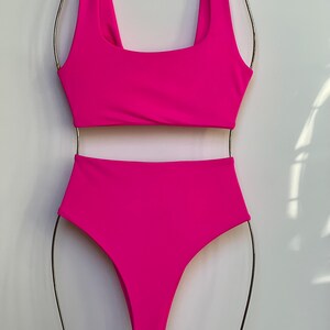 May include: A vibrant fuchsia two-piece swimsuit set. The top has a square neckline and the high-waisted bottoms offer full coverage. The swimsuit is displayed on a metal hanger against a plain white background.