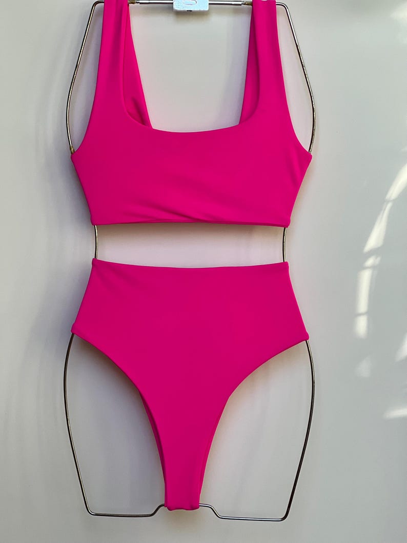 May include: A vibrant fuchsia two-piece swimsuit set. The top has a square neckline and the high-waisted bottoms offer full coverage. The swimsuit is displayed on a metal hanger against a plain white background.