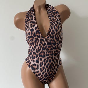 Puede incluir: Un traje de baño de una pieza con estampado de leopardo en tonos marrón y negro. La prenda presenta un escote halter con una parte delantera en forma de V profunda, un corpiño estilo cruzado y aberturas de pierna altas, sobre un torso de maniquí.