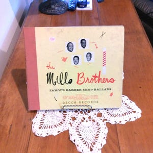 May include: A vintage vinyl record album titled "The Mills Brothers Famous Barber Shop Ballads" with a light tan cover and red spine. The cover features four black and white portraits and the Decca Records logo.