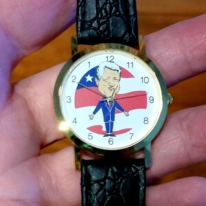 May include: A gold-toned wristwatch with a black leather band. The watch face features a cartoon illustration of a person in a blue suit with a red, white, and blue background. The watch has black numbers and hands.