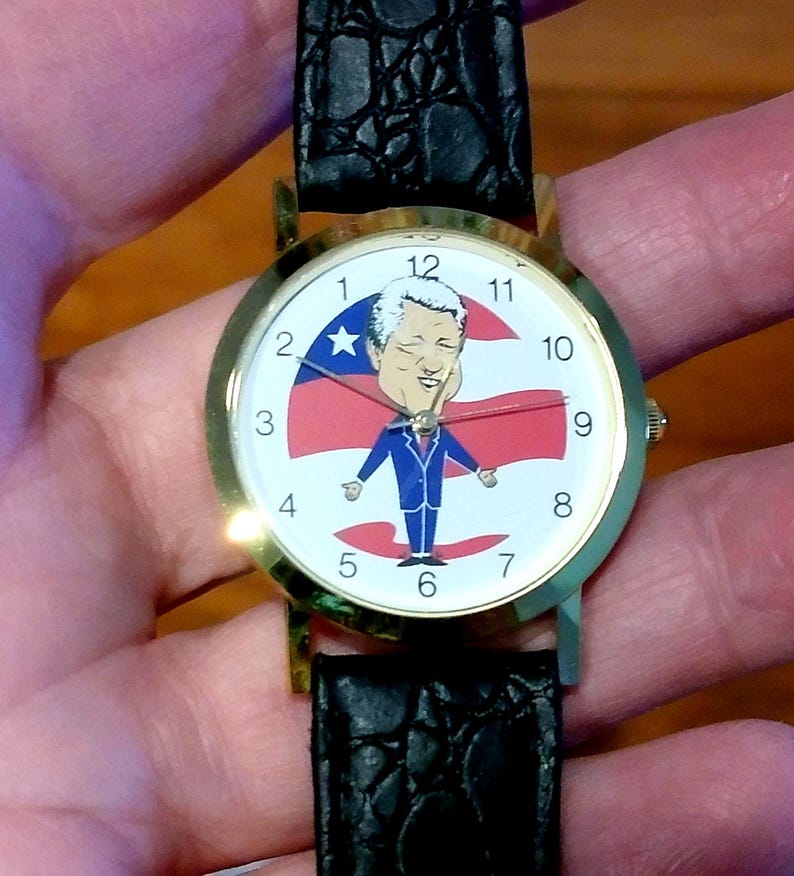 May include: A gold-toned wristwatch with a black leather band. The watch face features a cartoon illustration of a person in a blue suit with a red, white, and blue background. The watch has black numbers and hands.