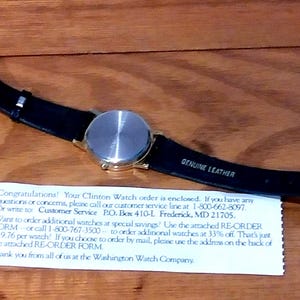 May include: A wristwatch with a gold-colored case and a black leather strap. The watch face is silver. The strap is labeled "GENUINE LEATHER". A white paper with text is below the watch.