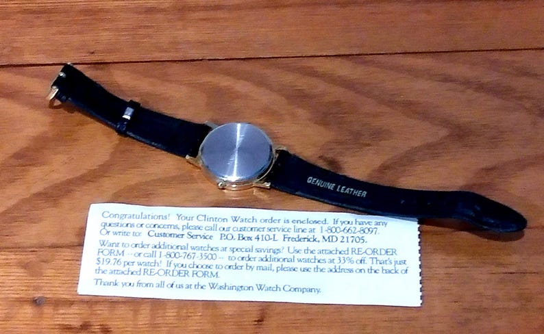 May include: A wristwatch with a gold-colored case and a black leather strap. The watch face is silver. The strap is labeled "GENUINE LEATHER". A white paper with text is below the watch.