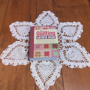 May include: A copy of "The Quilting Answer Book" rests on a white, star-shaped doily. The book has a red and pink cover with the title in large, gold letters. The doily is made of white lace and is placed on a wooden surface.
