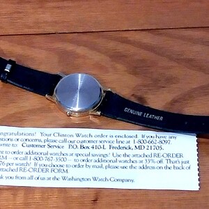 May include: A wristwatch with a silver-colored face and a black leather band. The band is labeled "GENUINE LEATHER." A white paper with text is beneath the watch. The watch is on a wooden surface.