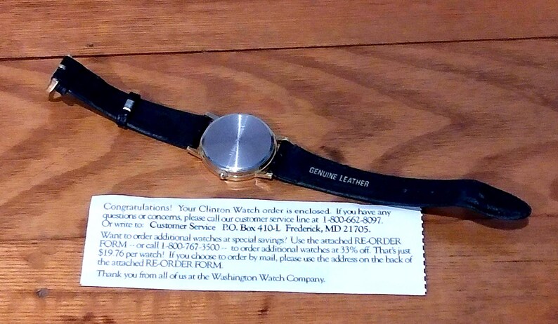 May include: A wristwatch with a silver-colored face and a black leather band. The band is labeled "GENUINE LEATHER." A white paper with text is beneath the watch. The watch is on a wooden surface.