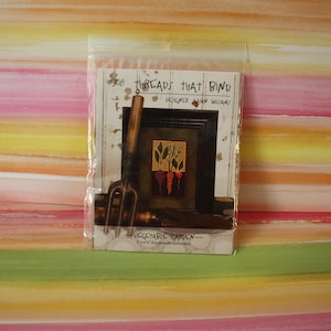 May include: A close-up of a packaged cross-stitch pattern titled "Vegetable Garden" by Threads That Bind, designed by Shawn Williams. The pattern features a small framed image of three carrots and a sprig of green leaves. The package also includes a small gardening tool.