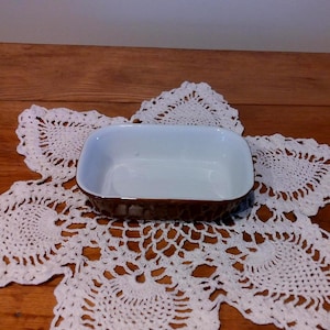 May include: A rectangular ceramic baking dish with a white interior and a brown textured exterior. The dish is set on a white lace doily, with a wooden surface in the background.
