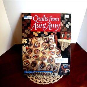 May include: A book titled "Quilts from Aunt Amy" with a red banner and white text. The cover features a quilt design with brown, tan, and blue circles. The book is on a dark wooden surface with a lace doily.