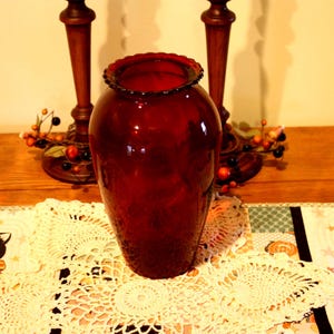 May include: A deep red glass vase with a ruffled rim. The vase is cylindrical with a rounded body. The background includes wooden candle holders and a decorative doily. Home decor.