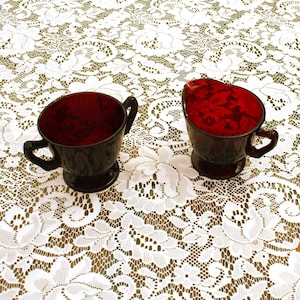 May include: Two dark red glass teacups with handles sit on a white lace tablecloth. One cup is upright, and the other is tilted, revealing its interior. The cups have a dark, textured appearance.