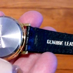 May include: A gold-toned wristwatch with a black leather strap. The watch has a silver-colored back and the strap is marked "GENUINE LEATHER". The watch appears to be new.