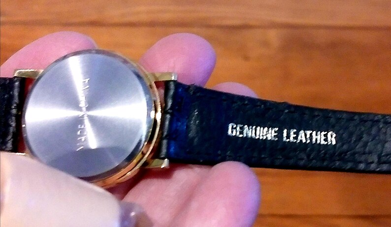 May include: A gold-toned wristwatch with a black leather strap. The watch has a silver-colored back and the strap is marked "GENUINE LEATHER". The watch appears to be new.