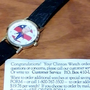 May include: A gold-toned wristwatch with a black leather band. The watch face features a patriotic design with red, white, and blue colors. The watch is displayed on a wooden surface.