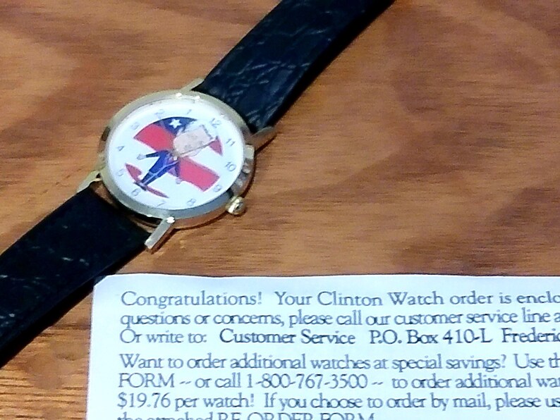May include: A gold-toned wristwatch with a black leather band. The watch face features a patriotic design with red, white, and blue colors. The watch is displayed on a wooden surface.