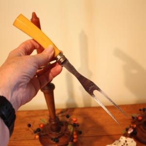 May include: A vintage carving fork with a yellow handle and a silver blade. The fork is held in a hand, with two wooden candle holders and candles in the background. The scene is set on a wooden surface.