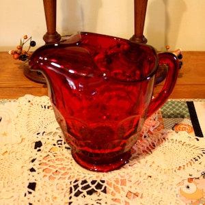 May include: A ruby red glass pitcher with a handle. The pitcher has a decorative pattern and a spout for pouring. It sits on a white lace doily and a patterned table runner. The background includes wooden elements and decorative accents.