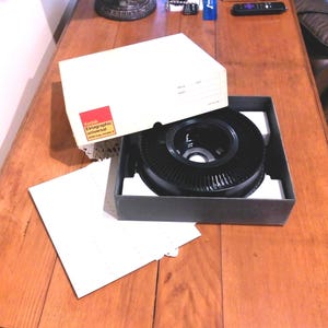 May include: A Kodak Ektagraphic Universal slide tray model 2 is displayed in its original box. The box is white with a red and yellow label. The slide tray is black and circular, with a ridged design. A white sheet of paper is next to the box.