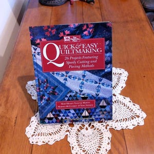 May include: A book titled "Quick & Easy Quiltmaking" with a red and white banner. The book features 26 projects with speedy cutting and piecing methods. It rests on a white, star-shaped doily on a wooden surface.