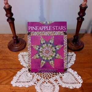 May include: A book titled "Pineapple Stars" with a quilt design on the cover. The cover features a star pattern in shades of pink, purple, and green. The book is on a white lace doily, with two wooden candle holders on either side.