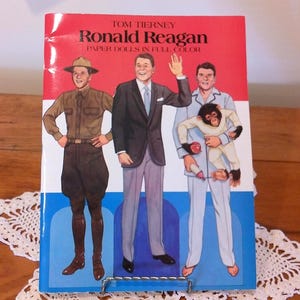 May include: A vintage book titled "Ronald Reagan Paper Dolls in Full Color" with illustrations of Ronald Reagan in different outfits. The cover is red, white, and blue, with the title in bold, black letters. The book is displayed on a metal stand.