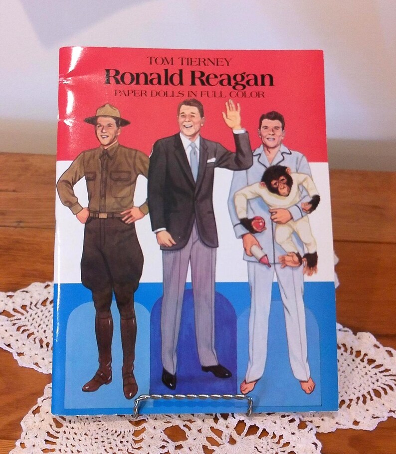 May include: A vintage book titled "Ronald Reagan Paper Dolls in Full Color" with illustrations of Ronald Reagan in different outfits. The cover is red, white, and blue, with the title in bold, black letters. The book is displayed on a metal stand.