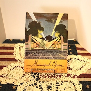 May include: A vintage Municipal Opera program from 1942, featuring a colorful illustration of an outdoor theater with spotlights and an audience. The cover has the words "Municipal Opera" in a stylized font, with additional text indicating the location and season.