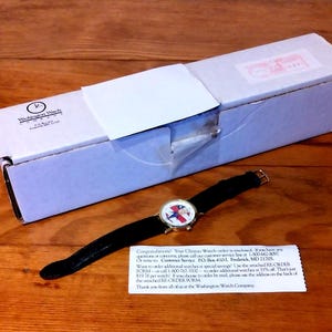 May include: A wristwatch with a black leather band and a gold-tone case, featuring a face with a patriotic design. The watch is displayed next to a white box with the text "Washington Watch" and a small note.