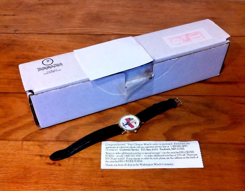 May include: A wristwatch with a black leather band and a gold-tone case, featuring a face with a patriotic design. The watch is displayed next to a white box with the text "Washington Watch" and a small note.