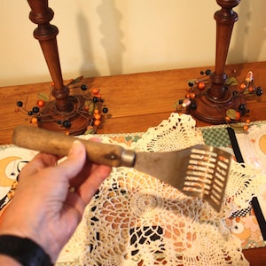 May include: An antique potato masher with a wooden handle and a slotted metal head. The masher is held over a cream-colored doily and a Halloween-themed table runner. Two wooden candle holders with berry wreaths are in the background.