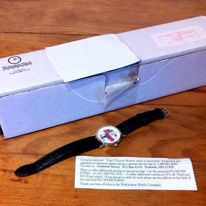 May include: A wristwatch with a black leather band and a gold-colored case. The watch face features a design with red, white, and blue colors. The watch is next to a white box with a logo and a small note.
