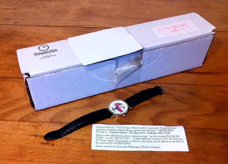 May include: A wristwatch with a black leather band and a gold-colored case. The watch face features a design with red, white, and blue colors. The watch is next to a white box with a logo and a small note.