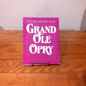 May include: A pink "Picture-History Book" titled "Grand Ole Opry" in large white letters. The book is displayed on a metal stand, set against a wooden surface.