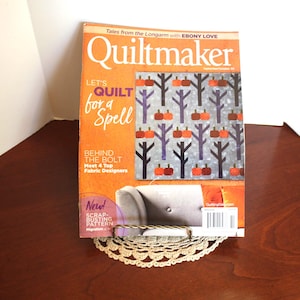 May include: A copy of Quiltmaker magazine with an orange cover featuring the text "Let's Quilt for a Spell." The cover art displays a quilt design with trees and pumpkins. The magazine rests on a decorative metal and lace stand.
