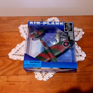 May include: A toy airplane in a clear plastic box. The airplane is painted in green and red camouflage with a red circle on the side. The box has the words "AIR-PLANE" printed on it.