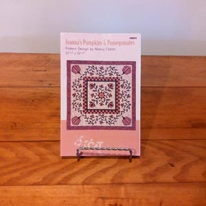 May include: A pattern design titled "Joanna's Pumpkins & Pomegranates" by Nancy Odom, displayed on a metal stand. The design features a floral and geometric pattern in shades of purple, red, and cream. The pattern measures 57" x 57".