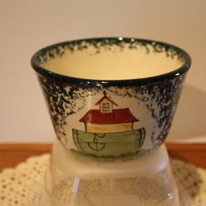 May include: A white ceramic bowl with a green and white speckled glaze. The bowl has a painted design of a green and white boat with a red roof and a white window. The boat has a green anchor on the side.
